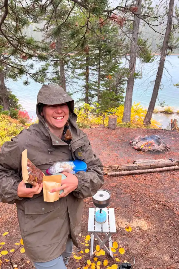 Mindy holding homemade freeze dried camping meals in a rain suit next to a propane camp stove and campfire