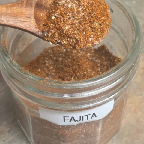 homemade fajita seasoning in a glass jar with a wooden spoon on a slate surface
