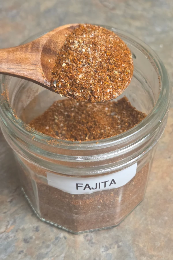 homemade fajita seasoning in a glass jar with a wooden spoon on a slate surface